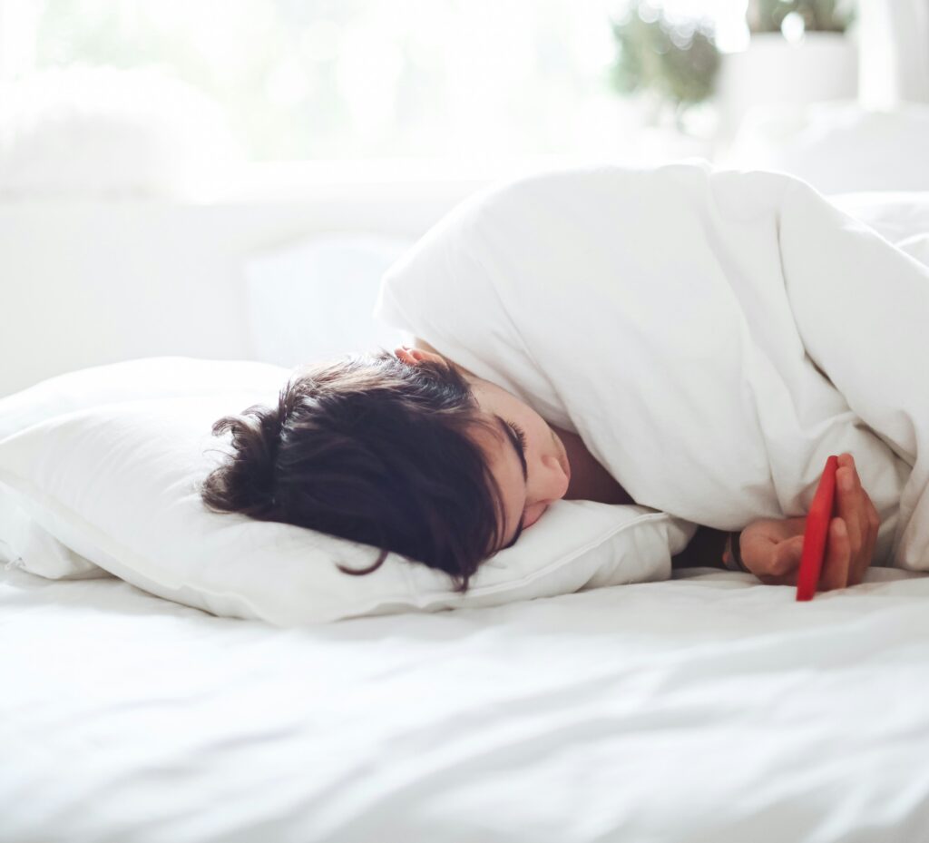 Frau schläft morgens im hellen Bett, die Bettdecke über dem Kopf und das Handy in der Hand – zu wenig Schlaf und Heißhunger am Folgetag