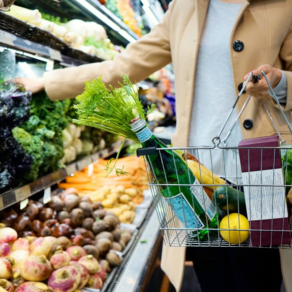 Gesund einkaufen mit wenig Zeit: Person greift in der Gemüseabteilung eines Supermarkts nach frischem Blattgemüse, in der anderen Hand ein Einkaufskorb mit Obst und Gemüse
